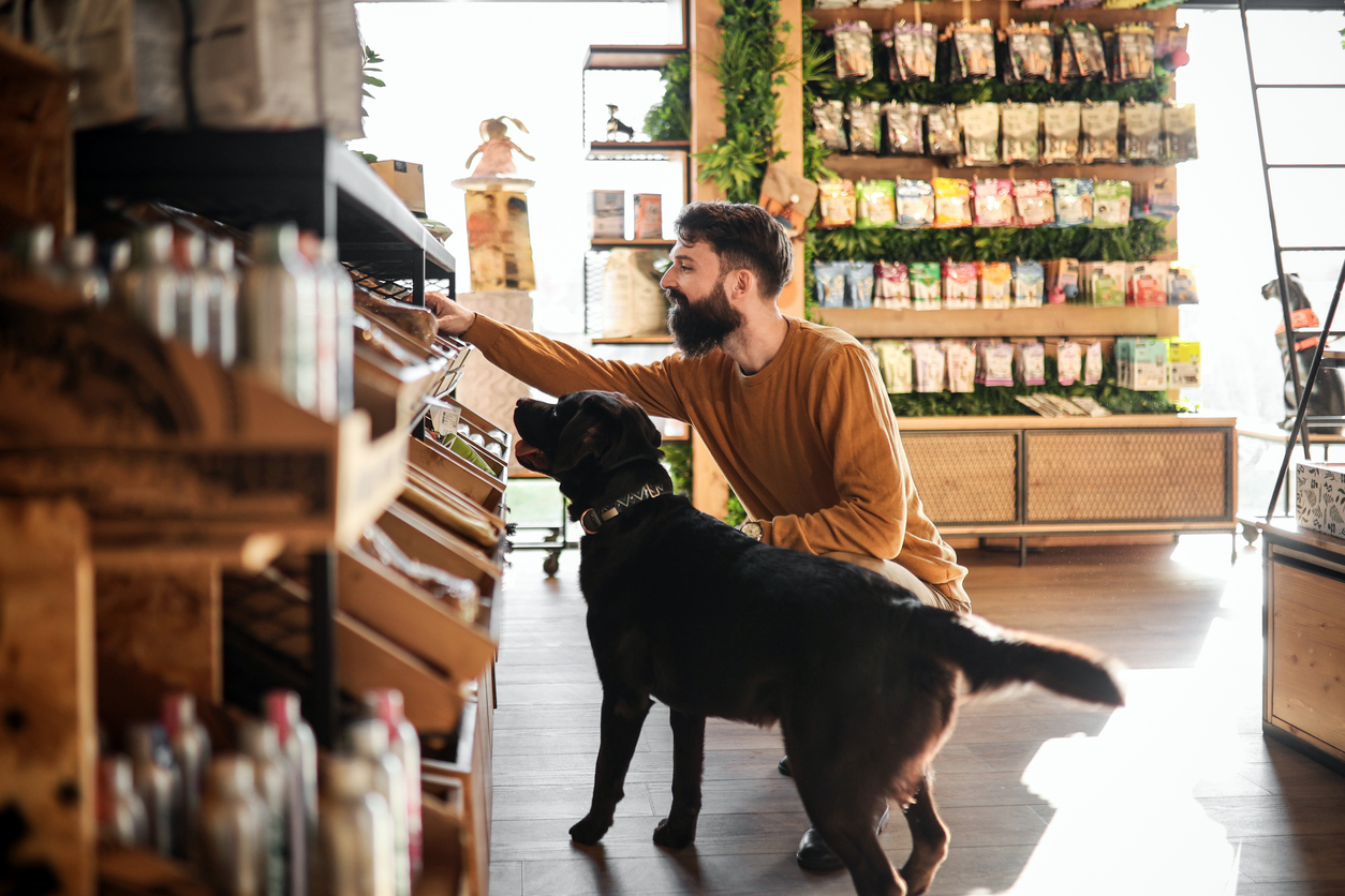 A bearded man in a mustard sweater crouches in a pet store aisle, reaching for an item on the shelf while his black Labrador stands beside him. The background shows rows of colorful pet food bags and greenery on the wall, with wooden shelves and crates neatly displaying products.